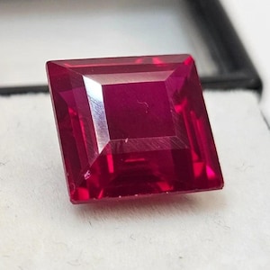 Può includere: Un rubino sfaccettato a taglio quadrato. La gemma è di un colore rosso intenso e saturo, con bordi netti e definiti. Il rubino è presentato su uno sfondo bianco, evidenziandone la brillantezza e la chiarezza.