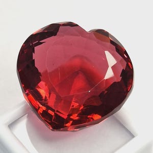 Pode incluir: Uma pedra preciosa facetada em forma de coração, em um tom vermelho profundo. A gema tem múltiplas facetas que refletem a luz, criando um efeito brilhante. A pedra preciosa está colocada sobre um fundo branco.