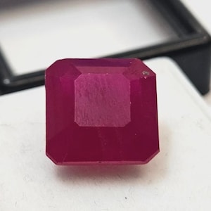 Puede incluir: Una gema de rubí tallada en forma cuadrada y facetada. La gema es de color rojo intenso y saturado, con forma rectangular y bordes biselados. El rubí se encuentra sobre una superficie blanca, con un marco negro en el fondo.