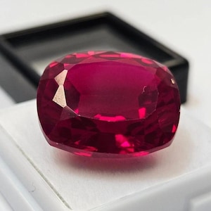 Op de afbeelding: Een gefacetteerde robijn edelsteen met kussensnede. De dieprode edelsteen wordt gepresenteerd op een wit oppervlak, met een zwarte doos op de achtergrond. De facetten van de robijn reflecteren licht, wat de glans en kleur laat zien.