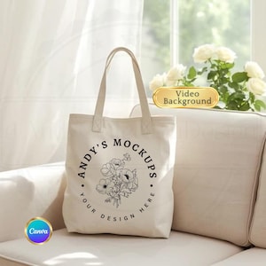 May include: Cream-colored tote bag with the text "ANDY'S MOCKUPS" encircling a floral design. The words "YOUR DESIGN HERE" are at the bottom of the circle. The bag has a simple design with two handles and is sitting on a cream-colored couch.