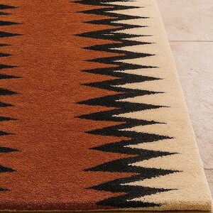 May include: A close-up of a rug with a geometric pattern. The rug features a zig-zag design in black against a backdrop of brown and beige. The rug's texture is visible, suggesting a soft, woven material. The rug is a home decor item.