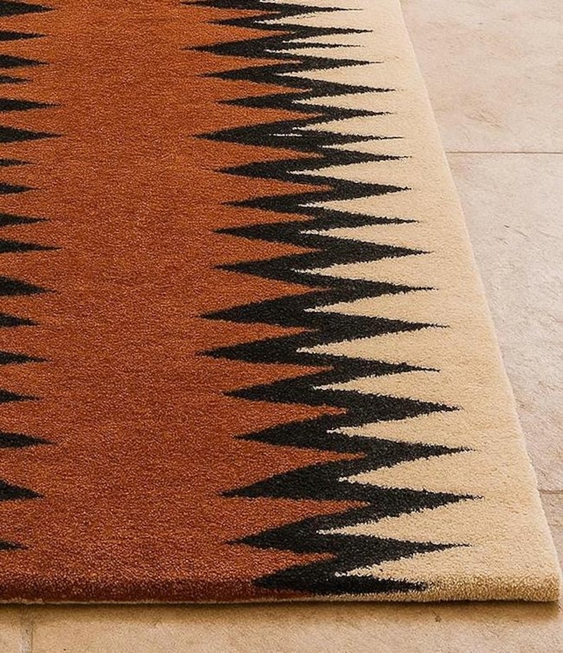 May include: A close-up of a rug with a geometric pattern. The rug features a zig-zag design in black against a backdrop of brown and beige. The rug's texture is visible, suggesting a soft, woven material. The rug is a home decor item.