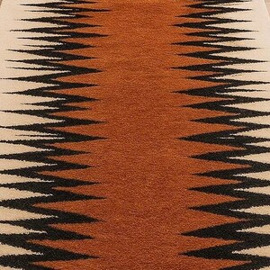 May include: A rectangular rug with a central band of orange-brown color, flanked by cream-colored borders. The rug features a repeating zigzag pattern in black, creating a striking visual contrast. The rug is likely made of a soft, textured material.