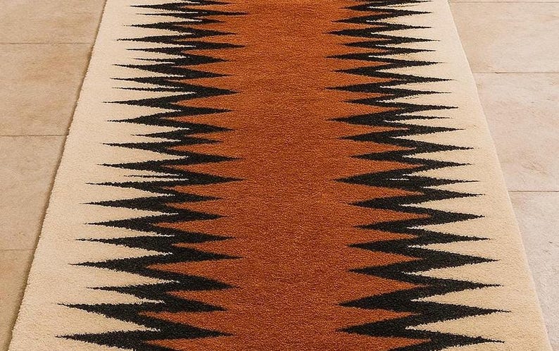 May include: A rectangular rug with a central band of orange-brown color, flanked by cream-colored borders. The rug features a repeating zigzag pattern in black, creating a striking visual contrast. The rug is likely made of a soft, textured material.