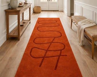 May include: An orange runner rug with a dark red abstract design. The rug is rectangular and placed in a hallway with light-colored walls and wooden flooring. The design is a continuous line.