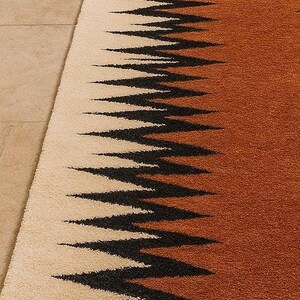 May include: A rectangular rug with a geometric design. The rug features a cream-colored section on the left, transitioning to a brown hue on the right. Black zigzag patterns run across the rug, creating a striking visual contrast.