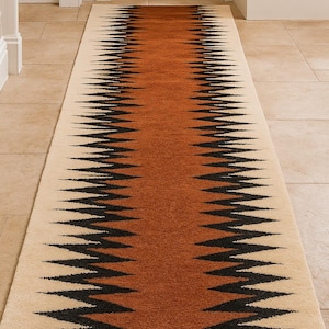 May include: A long, rectangular rug with a geometric pattern. The rug features a central band of brown, flanked by cream-colored borders. Black zigzag lines border the brown section, creating a striking visual contrast. The rug is placed in a hallway. in warm light