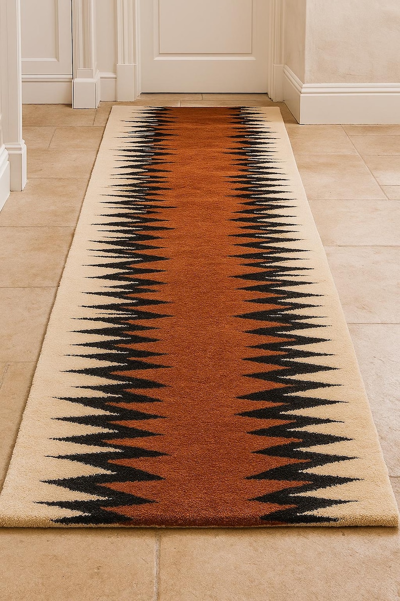 May include: A long, rectangular rug with a geometric pattern. The rug features a central band of brown, flanked by cream-colored borders. Black zigzag lines border the brown section, creating a striking visual contrast. The rug is placed in a hallway. in warm light