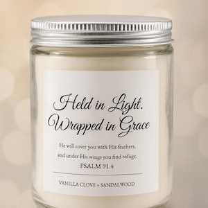 Puede incluir: Una vela en tarro de vidrio transparente con tapa plateada. La etiqueta dice "Held in Light. Wrapped in Grace" con un versículo bíblico y descripción de la fragancia. La vela tiene aroma a vainilla, clavo y sándalo.