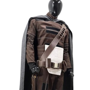 Puede incluir: Un disfraz marr&oacute;n y negro con una capa gris, exhibido en un maniqu&iacute;. El disfraz presenta un cintur&oacute;n de cuero marr&oacute;n, una secci&oacute;n media acolchada blanca y una bandolera con bolsillos. Las mangas tienen patrones geom&eacute;tricos.