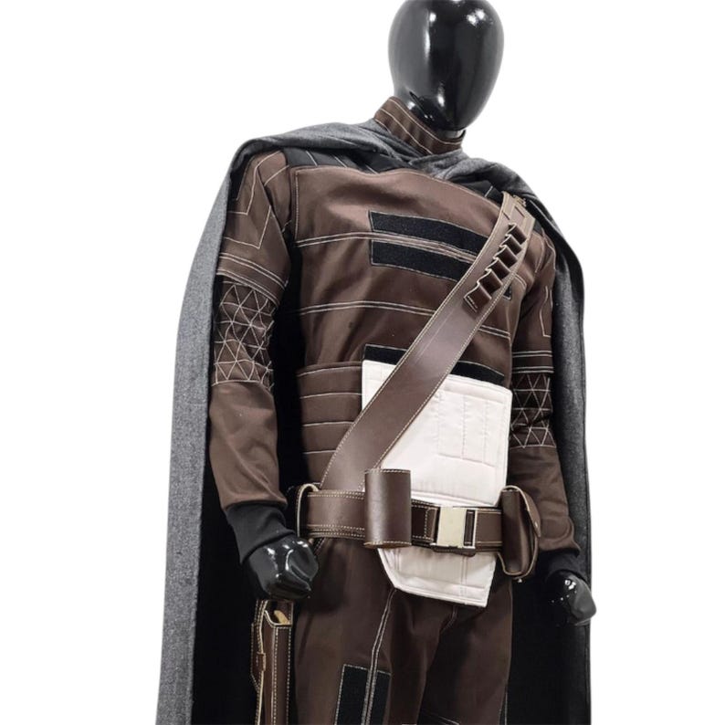 Puede incluir: Un disfraz marr&oacute;n y negro con una capa gris, exhibido en un maniqu&iacute;. El disfraz presenta un cintur&oacute;n de cuero marr&oacute;n, una secci&oacute;n media acolchada blanca y una bandolera con bolsillos. Las mangas tienen patrones geom&eacute;tricos.
