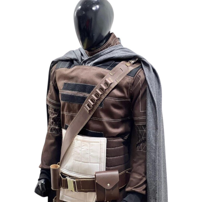 Puede incluir: Un disfraz marr&oacute;n y negro con una capa gris, exhibido en un maniqu&iacute;. El disfraz presenta una correa para el hombro con m&uacute;ltiples soportes cil&iacute;ndricos, un cintur&oacute;n con una bolsa y una pieza rectangular blanca en la parte delantera.
