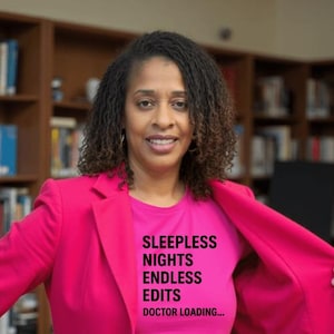 May include: A woman in a pink blazer and a pink t-shirt with the text "SLEEPLESS NIGHTS ENDLESS EDITS DOCTOR LOADING..." in black. The background shows bookshelves, suggesting a library or study setting.