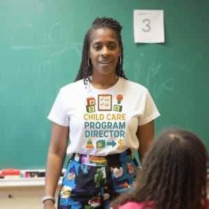 May include: A white t-shirt with the text "CHILD CARE PROGRAM DIRECTOR" in blue and orange. The shirt has colorful graphics of blocks, a checklist, and a lightbulb. The person is standing in front of a green chalkboard.