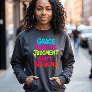 May include: Dark gray sweatshirt with the text "GRACE made me. JUDGMENT can't BREAK ME" in bright, dripping font. The sweatshirt has a kangaroo pocket and long sleeves. The person in the image is wearing the sweatshirt.