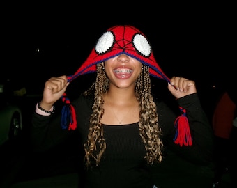 May include: A red, blue, and white Spider-Man knit hat with white eye details. The hat has long ear flaps with red and blue tassels. The person is wearing a black long-sleeve shirt.