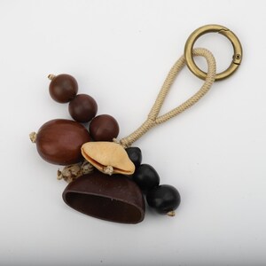 Op de afbeelding: Een handgemaakte sleutelhanger met een verzameling natuurlijke elementen. Het bevat diverse bruine, zwarte en beige zaden en peulen, samen met een goudkleurige metalen sluiting en een beige touwlus. De sleutelhanger is ontworpen om aan sleutels of tassen te bevestigen.