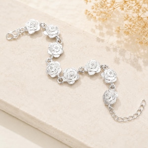 May include: A silver-toned bracelet featuring seven repeating flower-shaped links connected by small circular rings. The bracelet includes a lobster claw clasp at one end and an adjustable chain extension with several links at the opposite end.