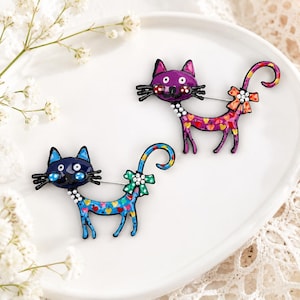 May include: Two cat-shaped enamel pins, one purple and one blue, each featuring a colorful heart pattern on the body, a small bow with embedded rhinestones at the tail, and a row of white faux pearls along the neck area with black whiskers.