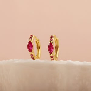 May include: A pair of gold hoop earrings. Each earring has a central, marquise-cut ruby gemstone, with smaller ruby accents. The earrings are presented on a white, textured surface against a soft pink backdrop.