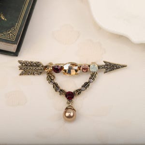 Peut inclure: Broche dorée antique en forme de flèche, ornée de pierres précieuses colorées et d'une perle pendante. La broche présente une pierre centrale ovale, entourée de petites pierres de couleurs rouge, rose et transparent. Mesure environ 7 cm de long.