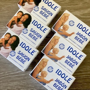 Könnte beinhalten: Mehrere weiße Schachteln mit IDOLE Savon Bébé Seife liegen auf einer Holzoberfläche. Die Schachteln zeigen Bilder von Mutter und Kind sowie den Text "IDOLE SAVON BÉBÉ" in Blau. Die Seife enthält Glycerin und Vitamin E.