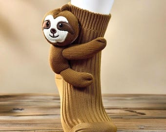May include: A brown ribbed crew sock featuring a plush, three-dimensional sloth attached to the side. The sloth has a white face, dark brown eye patches, and embroidered facial features, with its arms wrapped around the sock fabric.