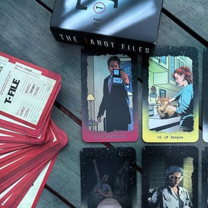 Könnte beinhalten: Ein Tarotkartenspiel mit Illustrationen, die von einer Fernsehserie inspiriert sind, auf einer Holzoberfläche. Die Karten zeigen verschiedene Charaktere und Szenen mit Titeln wie "Der Teufel" und "Die Partner". Eine Schachtel mit der Aufschrift "THE TAROT FILES" ist ebenfalls vorhanden.