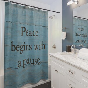 May include: A blue shower curtain with the words "Peace begins with a pause" in dark brown lettering. The curtain has a wood-grain design and is hanging in a bathroom with a white vanity and a mirror.