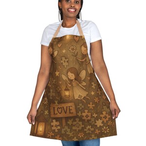May include: Brown apron with a whimsical fairy design. The apron features illustrations of fairies, flowers, puzzle pieces, and the word "LOVE" on a wooden sign. The apron has a light brown neck strap and waist ties.
