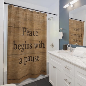 May include: A brown shower curtain with the text "Peace begins with a pause" in black. The curtain has a wood grain design. The bathroom features white cabinets, a marble countertop, and a dark grey tiled floor.