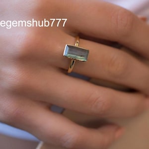 May include: A gold ring with a rectangular, iridescent gemstone. The ring has a thin, polished gold band. The gemstone shows shades of blue, green, and grey. The text "Thegemshub777" is visible in the upper left corner.