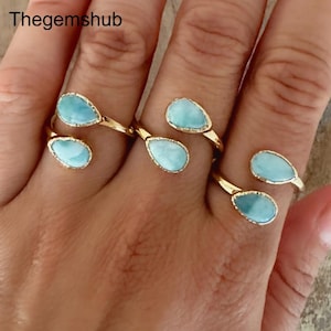 May include: Gold-toned rings with teardrop-shaped, light blue gemstones. The rings are open-ended, with a gemstone at each end. The rings are displayed on a hand, with the shop name "Thegemshub" visible in the upper left corner.
