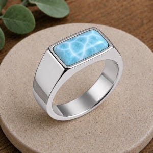May include: A silver-toned ring featuring a rectangular, light blue stone with a white, web-like pattern. The band is smooth and polished, with a flat, wide top setting that holds the stone securely in place, creating a clean and solid appearance.