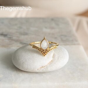 May include: A gold ring featuring a pear-shaped, white gemstone. The ring has a V-shaped design with small, beaded details. The ring is presented on a white, oval stone. The text "Thegemshub" is visible in the top left corner.