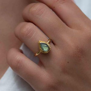 Peut inclure: Bague dorée ornée d'une pierre précieuse irisée en forme de goutte. La bague présente un bord délicat et perlé autour de la pierre et un anneau fin. Thegemsub777 est visible.