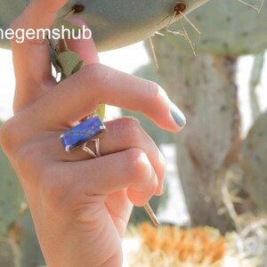 May include: A silver ring featuring a rectangular blue gemstone. The gemstone displays a marbled pattern with blue and purple hues. The ring is set against a blurred backdrop of green plants. The text "Thegemshub" is visible in the image.