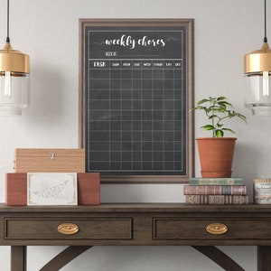 May include: A wooden table with two drawers and a chalkboard with the text "weekly chores" and a grid for writing tasks. There are two gold pendant lights with clear glass shades hanging above the table. A potted plant, a stack of books, and a wooden box with a label that says "The Avocado Toast" are on the table.