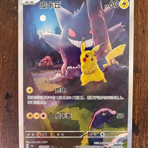 Puede incluir: Una carta coleccionable de Pokémon con Pikachu y Gengar en un bosque. La tarjeta tiene texto e ilustraciones en japonés, con un Pikachu amarillo en primer plano y un Gengar morado en el fondo. La tarjeta tiene 60 HP.