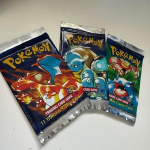 Puede incluir: Tres paquetes sellados de cartas coleccionables Pokémon. Los paquetes presentan imágenes de Charizard, Blastoise y otros personajes Pokémon. Cada paquete es azul marino con bordes de lámina plateada y el logotipo de Pokémon.