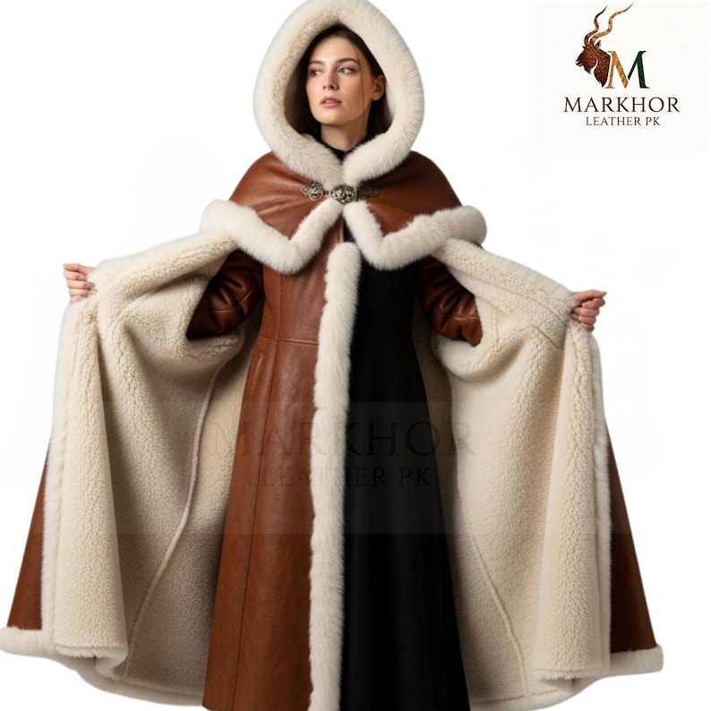 May include: A brown leather hooded cloak with a cream-colored fur trim and lining. The cloak features a decorative clasp at the neckline and is open, revealing a black dress. The Markhor Leather PK logo is in the upper right corner.