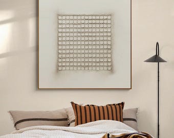 Könnte beinhalten: Quadratische abstrakte Kunst mit einem Rastermuster in einer neutralen Farbpalette. Das Kunstwerk ist in einem dünnen, braunen Holzrahmen gerahmt und hängt über einem Bett mit dekorativen Kissen und einer Decke. Rechts befindet sich eine schwarze Stehlampe.