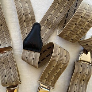 Könnte beinhalten: Beige und schwarz gemusterte Hosenträger mit silberfarbenen Metallverschlüssen. Die Hosenträger haben einen schwarzen Leder-Rückenaufnäher mit Monogramm. Der Stoff hat ein gewebtes Design mit vertikalen Streifen und kleinen schwarzen Quadraten.