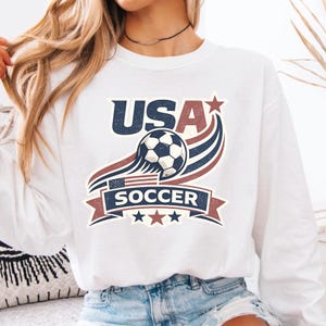 Peut inclure: Un sweat-shirt blanc &agrave; manches longues avec un motif graphique portant le texte USA SOCCER. Le design comprend un ballon de football, des rayures courbes rouges et bleues, un petit drapeau am&eacute;ricain et trois &eacute;toiles dans un style vintage.
