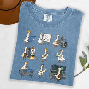 May include: Blue t-shirt featuring a whimsical design of cartoon geese in various professions, including an engineer, scientist, and technician. The geese are depicted with tools and equipment, such as blueprints, a rocket, and a computer. The shirt has a crew neck and short sleeves.