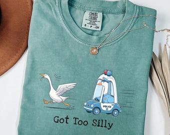 May include: Teal Comfort Colors t-shirt with a whimsical design. A white goose runs from a blue police car driven by a goose. The text "Got Too Silly" is printed below the graphic. A silver necklace is draped across the shirt.