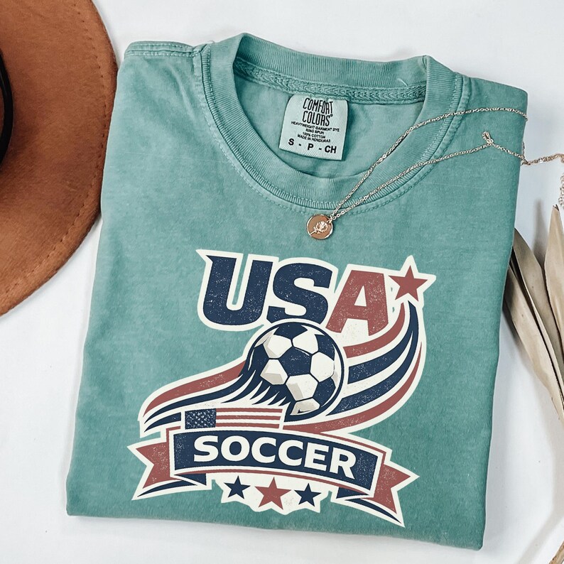 Peut inclure: Un t-shirt &agrave; manches courtes vert sauge avec une impression graphique portant le texte USA SOCCER. Le motif comprend un ballon de football, un petit drapeau am&eacute;ricain et des rayures rouges, blanches et bleues avec des &eacute;toiles. &Eacute;tiquette Comfort Colors.