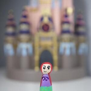 May include: A hand-painted wooden peg doll with a red hood, purple top, and turquoise skirt with a green pattern. The doll has a smiling face and is in front of a blurred pink and blue castle.