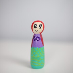 May include: A hand-painted wooden peg doll in the shape of a mermaid. The doll features a red head, a purple top, and a turquoise skirt with a green scale pattern. It is adorned with purple gems and has a smiling face.